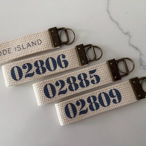 May include: Four canvas keychains with dark blue text. One keychain reads "RHODE ISLAND", the others display the numbers "02806", "02885", and "02809". Each keychain has a bronze-colored metal clasp.