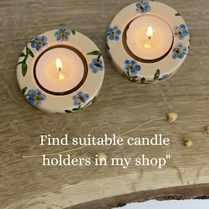 May include: Two cream-colored candle holders with blue floral designs and lit candles. The candle holders have a circular shape with a central opening for the candle. The text "Find suitable candle holders in my shop" is visible.