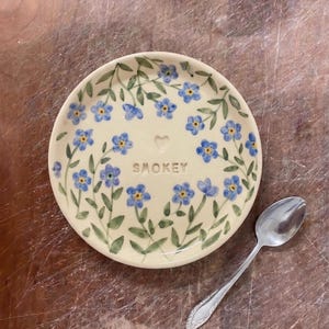 May include: A cream-colored ceramic plate with a floral design of blue flowers and green leaves. The word "SMOKEY" is written in the center. A silver spoon rests on the side of the plate.