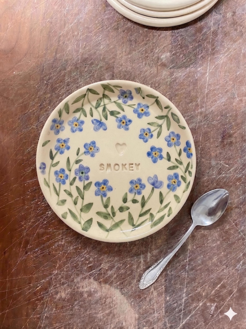 May include: A cream-colored ceramic plate with a floral design of blue flowers and green leaves. The word "SMOKEY" is written in the center. A silver spoon rests on the side of the plate.