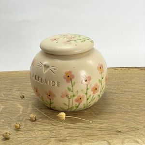 Op de afbeelding: Een kleine witte keramische pot met een deksel, versierd met roze bloemen en groene stelen. De pot heeft de naam "Adelaide" erop geschreven en een kattengezicht met een hart.