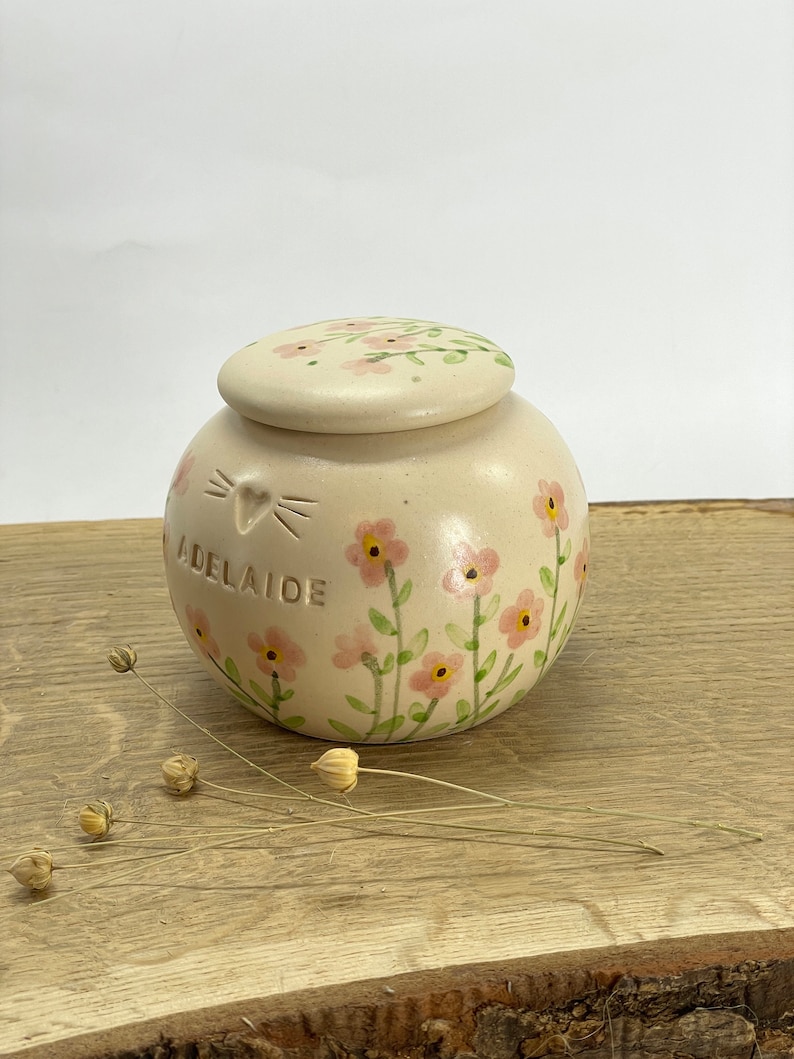 Op de afbeelding: Een kleine witte keramische pot met een deksel, versierd met roze bloemen en groene stelen. De pot heeft de naam "Adelaide" erop geschreven en een kattengezicht met een hart.