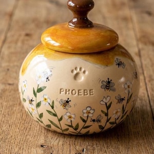 May include: A ceramic pet treat jar with a lid. The jar is beige with a paw print and the name "PHOEBE" in embossed letters. It is decorated with painted bees and flowers. The lid and handle are brown and yellow.