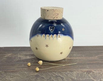 May include: A blue and white ceramic jar with a cork lid. The jar has a heart and the name "Chara" written on it. The jar is decorated with gold stars.