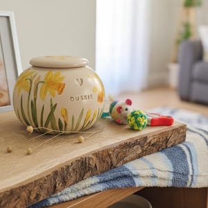 Pode incluir: Uma urna para animais de estimação creme com tampa, adornada com flores amarelas de narcisos e caules verdes. A urna tem um recorte em forma de coração e o nome "DUSKIE" impresso. Um retrato de gato emoldurado e brinquedos para gatos estão por perto.