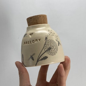 Könnte beinhalten: Eine weiße Keramikdose mit einem Korkdeckel, verziert mit einem Löwenzahnmuster und dem Wort "MALLORY" auf der Vorderseite. Die Dose hat ein Herzsymbol über dem Namen.