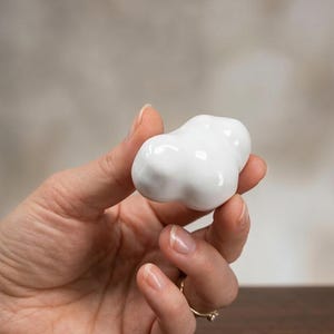 Può includere: Un piccolo oggetto in ceramica bianca a forma di nuvola tenuto in mano. L'oggetto ha una finitura liscia e lucida e una forma arrotondata e organica. Lo sfondo è di un tono neutro e sfocato.