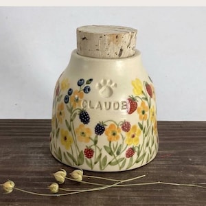 Könnte beinhalten: Ein cremefarbener Keramikbehälter mit Korkdeckel, verziert mit einem handgemalten Blumenmuster aus gelben Blüten, roten Erdbeeren und dunklen Beeren. Der Name CLAUDE ist vorne über einem Pfotenabdruck eingeprägt.