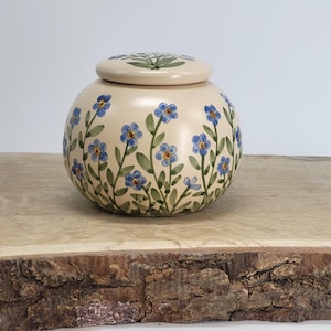 May include: A round, cream-colored ceramic jar with a matching lid. The jar and lid are decorated with blue forget-me-not flowers and green leaves. The jar is sitting on a wooden surface.