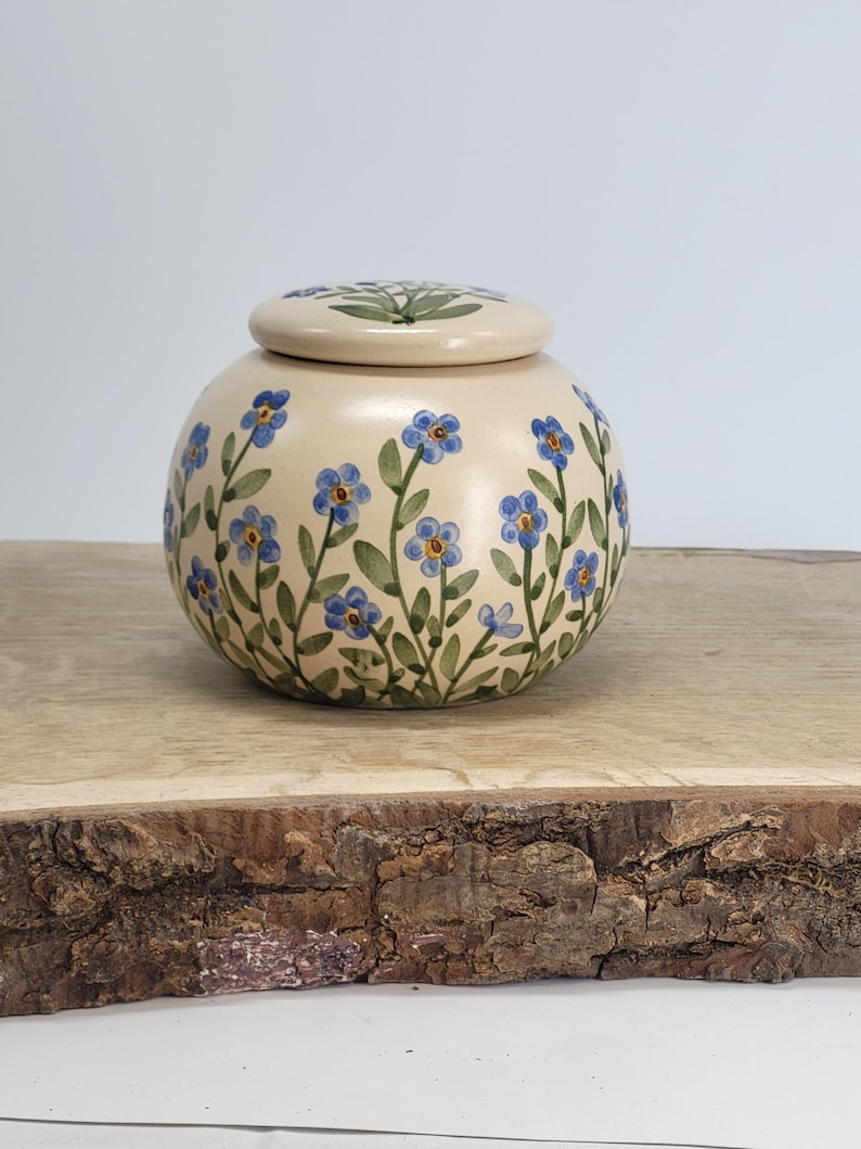 May include: A round, cream-colored ceramic jar with a matching lid. The jar and lid are decorated with blue forget-me-not flowers and green leaves. The jar is sitting on a wooden surface.