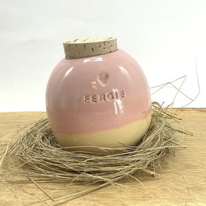 May include: A pink and cream ceramic egg-shaped jar with a cork lid. The jar has a heart and the name "Fergie" written on it. The jar is sitting in a nest of straw on a wooden surface.