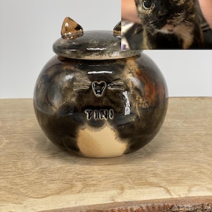 May include: A ceramic pet urn with a cat design. The round urn has a lid with cat ears. The body is a mix of brown, black, and tan colours, with the name "TINO" inscribed on the front. A photo of a cat is in the background.