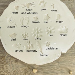Op de afbeelding: Witte keramische plaat met verschillende vormen en ontwerpen, waaronder een hart, een poot afdruk, een kruis, een kroon, een ster, een maan, een klavertjevier, vleugels, een bij, een wolk, een vogel, de zon, een madeliefje, een spruit, een vlinder, een veer en een davidster. Elke vorm is gelabeld met zijn naam.