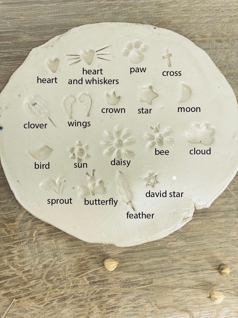 Op de afbeelding: Witte keramische plaat met verschillende vormen en ontwerpen, waaronder een hart, een poot afdruk, een kruis, een kroon, een ster, een maan, een klavertjevier, vleugels, een bij, een wolk, een vogel, de zon, een madeliefje, een spruit, een vlinder, een veer en een davidster. Elke vorm is gelabeld met zijn naam.