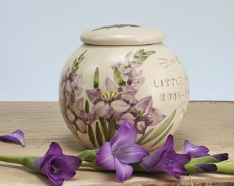 Può includere: Un'urna in ceramica color crema con coperchio, decorata con motivi floreali viola e foglie verdi. L'urna reca l'iscrizione "LITTLE ... 2005-". Fiori di gladiolo viola sono disposti attorno all'urna su una superficie di legno.