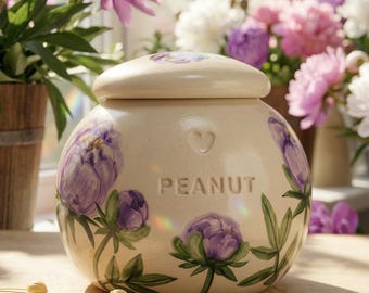 Può includere: Un vaso in ceramica color crema con coperchio, decorato con peonie viola dipinte a mano e foglie verdi. La parola "PEANUT" è in rilievo sul vaso. Una forma di cuore è scolpita vicino alla parte superiore. Il vaso è su una superficie di legno.