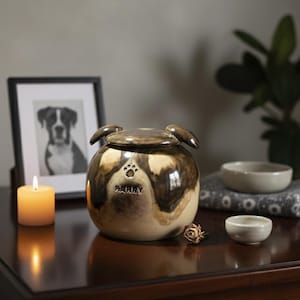 May include: A ceramic pet urn with a dog face design, featuring a paw print and the name "Barry." The urn is brown and cream-colored, with a lid and ear-shaped handles. A framed photo, candle, and bowls are nearby.