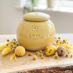 May include: A yellow ceramic jar with a lid, featuring the name "LOKI" and a sun design. The jar is surrounded by yellow decorative elements, including a crochet ball, a felt mouse, and a bell.