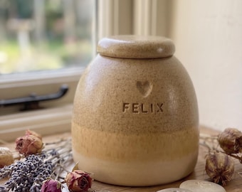 May include: A beige ceramic pet urn with a lid, featuring the name "FELIX" and a heart-shaped detail. The urn has a two-tone design and is surrounded by dried flowers and decorative stones with paw prints. A small, round locket is also present.