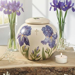 May include: Cream ceramic urn with a lid, decorated with large purple iris flower illustrations and a cross. The urn is on a wooden surface, with purple irises in vases and a lit candle. A decorative item.