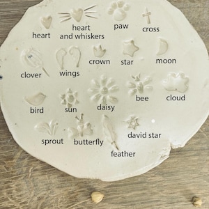 May include: A white, irregularly shaped ceramic piece with various embossed designs. The designs include a heart, paw print, cross, star, moon, clover, wings, bee, cloud, bird, sun, daisy, David star, sprout, butterfly, and feather. Text labels identify each design.