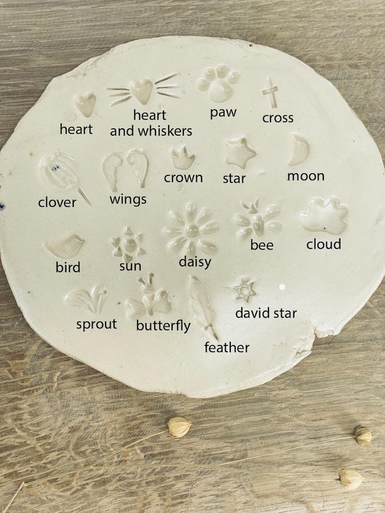 May include: A white, irregularly shaped ceramic piece with various embossed designs. The designs include a heart, paw print, cross, star, moon, clover, wings, bee, cloud, bird, sun, daisy, David star, sprout, butterfly, and feather. Text labels identify each design.