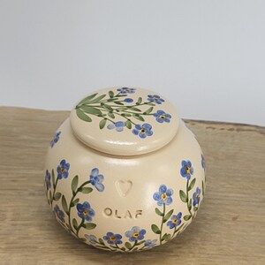 May include: A cream-colored ceramic jar with a lid, adorned with hand-painted blue forget-me-not flowers and green leaves. The jar has the name "OLAF" and a heart shape. The lid also features a floral design.
