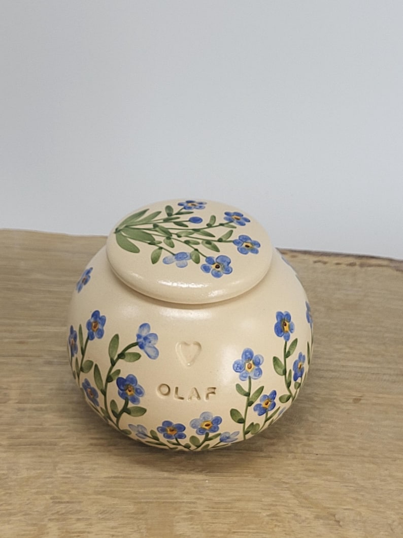 May include: A cream-colored ceramic jar with a lid, adorned with hand-painted blue forget-me-not flowers and green leaves. The jar has the name "OLAF" and a heart shape. The lid also features a floral design.