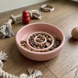 May include: A pink ceramic slow feeder dog bowl filled with kibble, alongside a dog toy, a rope toy, a collar, and a rubber ball. The bowl features a spiral design to slow down eating. The toys are red, beige, and orange.