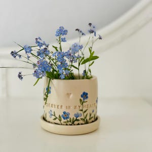Op de afbeelding: Een kleine, crèmekleurige keramische pot met een bijpassende schotel, gevuld met delicate blauwe vergeet-mij-nietjes. De pot is versierd met blauwe bloemmotieven en de woorden "FOREVER IN MY HEART". Een klein hartje is in de pot geëtst.