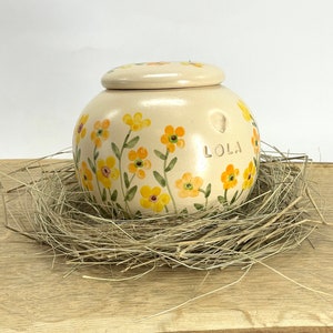 Op de afbeelding: Een crèmekleurige keramische pot met een deksel, versierd met gele en groene bloemen. De pot heeft de naam "LOLA" erop geschreven.