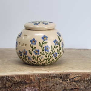 May include: A cream-colored ceramic jar with a lid, adorned with hand-painted blue forget-me-not flowers and green leaves. The jar has the word "CLAF" on the side. The jar is sitting on a wooden surface.