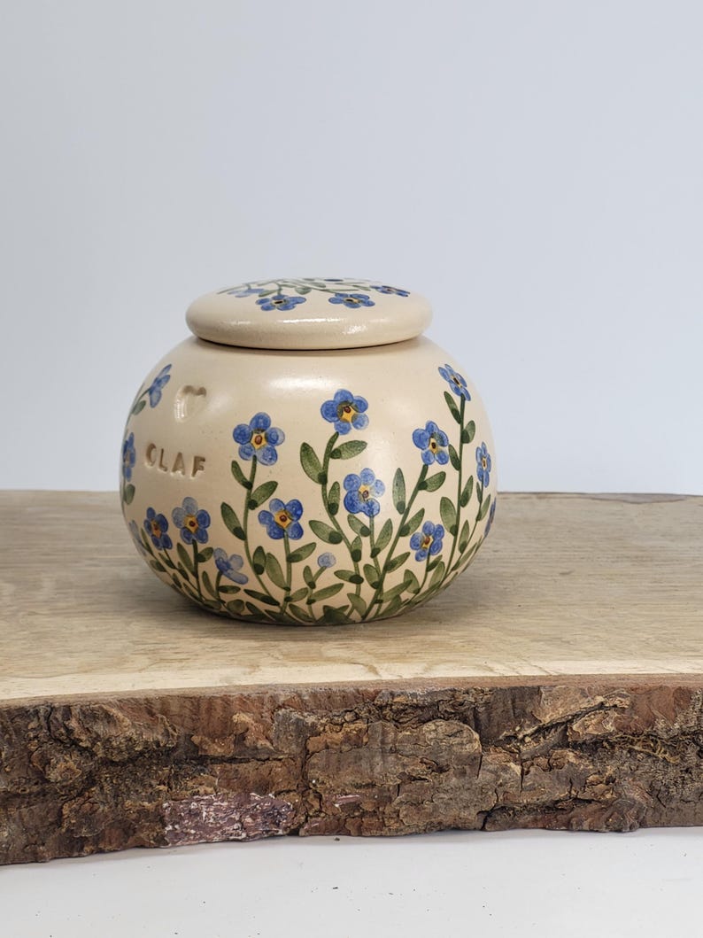 May include: A cream-colored ceramic jar with a lid, adorned with hand-painted blue forget-me-not flowers and green leaves. The jar has the word "CLAF" on the side. The jar is sitting on a wooden surface.