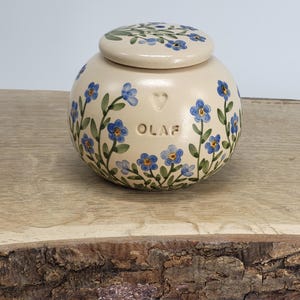 May include: A cream-colored ceramic jar with a lid, adorned with hand-painted blue forget-me-not flowers and green leaves. The jar has the name "OLAF" and a heart symbol. The lid is also decorated with the same floral pattern. The jar is sitting on a wooden surface.
