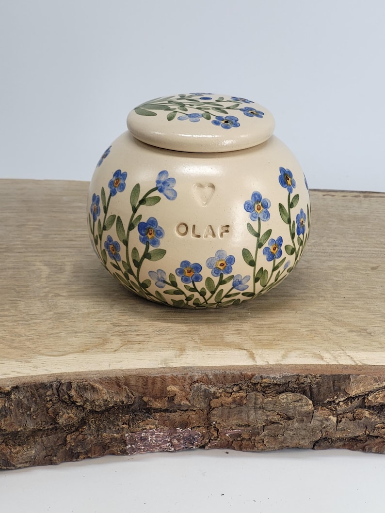 May include: A cream-colored ceramic jar with a lid, adorned with hand-painted blue forget-me-not flowers and green leaves. The jar has the name "OLAF" and a heart symbol. The lid is also decorated with the same floral pattern. The jar is sitting on a wooden surface.