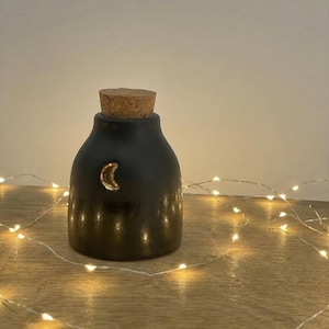 Puede incluir: Un tarro de cerámica negro con un tapón de corcho y un detalle de media luna dorada. El tarro está sobre una superficie de madera, con una guirnalda de pequeñas luces blancas cálidas enrollada alrededor. El tarro mide unos 10 cm de alto.
