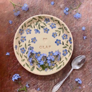 Könnte beinhalten: Ein cremefarbener Keramikteller mit einem Blumendesign aus blauen Vergissmeinnicht und grünen Blättern. Das Wort "OLAF" ist auf dem Teller geschrieben. Ein silberner Löffel und frische blaue Blumen liegen daneben.