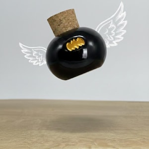 Pode incluir: Uma garrafa preta e redonda com rolha de cortiça e um emblema dourado. A garrafa parece estar flutuando, com asas de anjo brancas desenhadas atrás. A garrafa está em uma superfície de madeira.