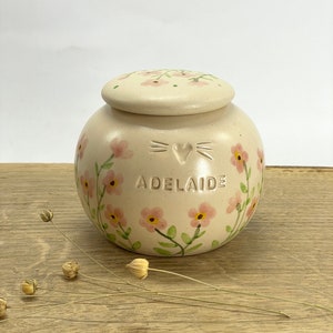 Op de afbeelding: Een cr&egrave;mekleurige keramische pot met een deksel, versierd met roze bloemen en groene bladeren. De pot heeft een hart en snorharen op de voorkant en de naam "Adelaide" eronder.