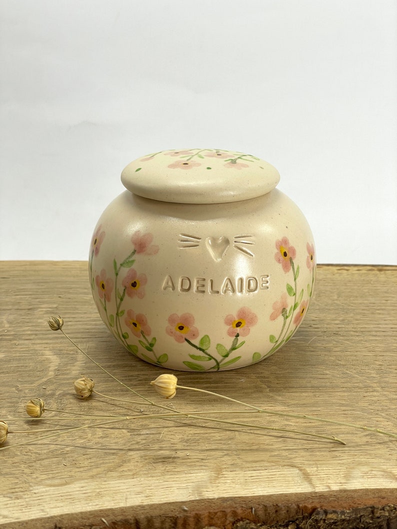 Op de afbeelding: Een cr&egrave;mekleurige keramische pot met een deksel, versierd met roze bloemen en groene bladeren. De pot heeft een hart en snorharen op de voorkant en de naam "Adelaide" eronder.