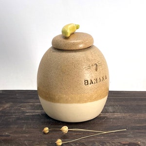 May include: A ceramic jar with a light brown glaze and a white band around the bottom. The jar has a banana-shaped lid and the word "Banana" is written on the front.