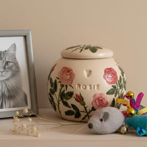 May include: A cream-colored ceramic pet urn with a lid, adorned with pink roses and green leaves. The name "ROSIE" is inscribed below a heart-shaped cutout. A framed black and white photo of a cat, a felt mouse toy, and a cat toy with bells are nearby.