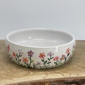 Puede incluir: Un cuenco redondo de cerámica blanca con un patrón floral pintado a mano en el exterior. El diseño incluye pequeñas flores rosas, moradas y lavanda claro con tallos y hojas verdes dispuestas en un motivo repetitivo alrededor del lateral.