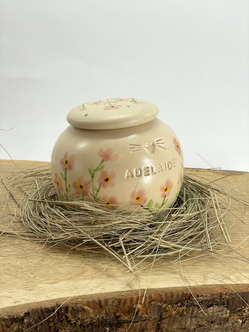 Op de afbeelding: Een kleine, cr&egrave;mekleurige keramische pot met een deksel, versierd met roze bloemen en de naam "Adelaide". De pot staat in een nest van stro op een houten oppervlak.