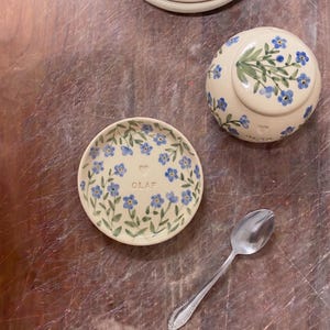 May include: A collection of cream-colored ceramic tableware with a floral design. The set includes small plates, a bowl, and a spoon. The plates and bowl are decorated with blue flowers and green leaves. The word "OLAF" is written on one of the plates.