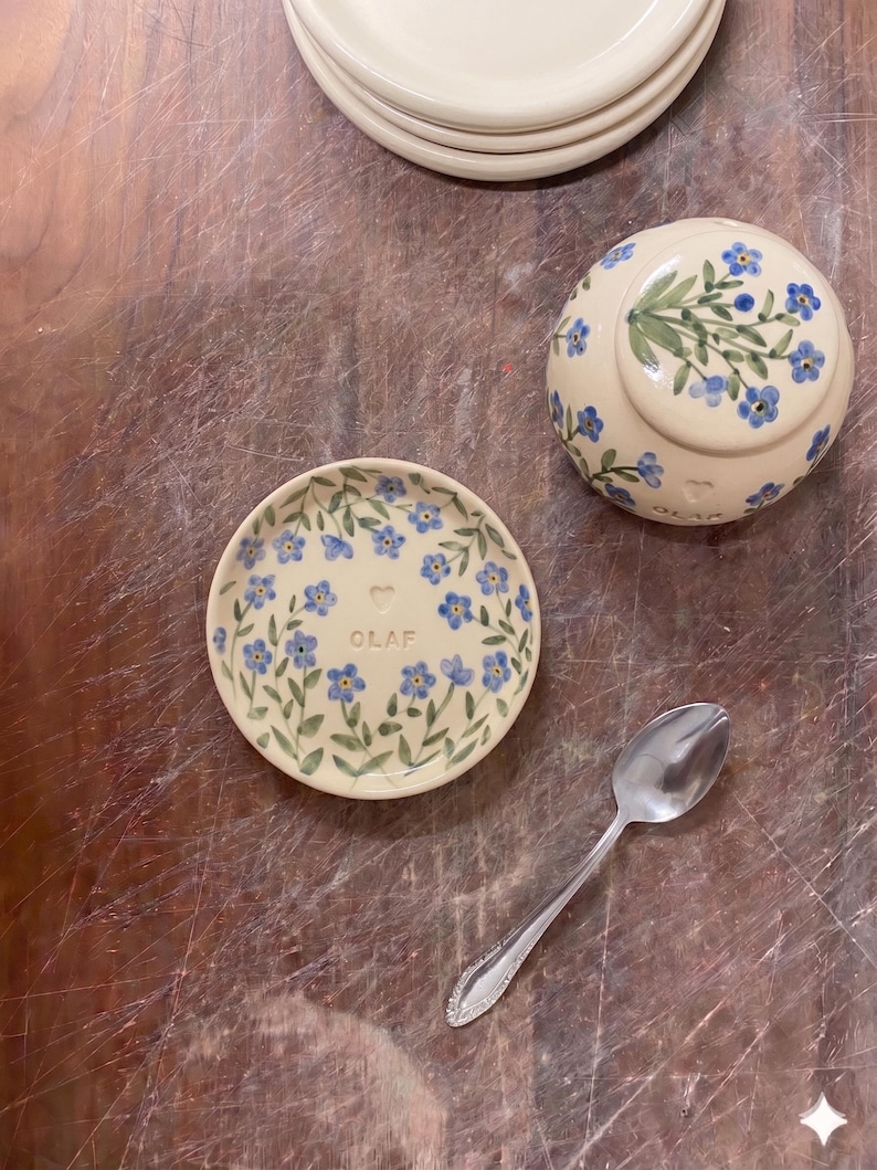 May include: A collection of cream-colored ceramic tableware with a floral design. The set includes small plates, a bowl, and a spoon. The plates and bowl are decorated with blue flowers and green leaves. The word "OLAF" is written on one of the plates.