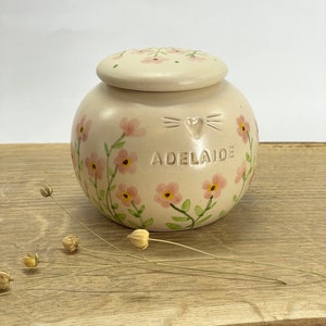 Op de afbeelding: Een kleine, cr&egrave;mekleurige keramische pot met een deksel, versierd met roze bloemen en groene bladeren. De pot heeft een kattengezicht en een hartontwerp en de naam "Adelaide" erop geschreven.