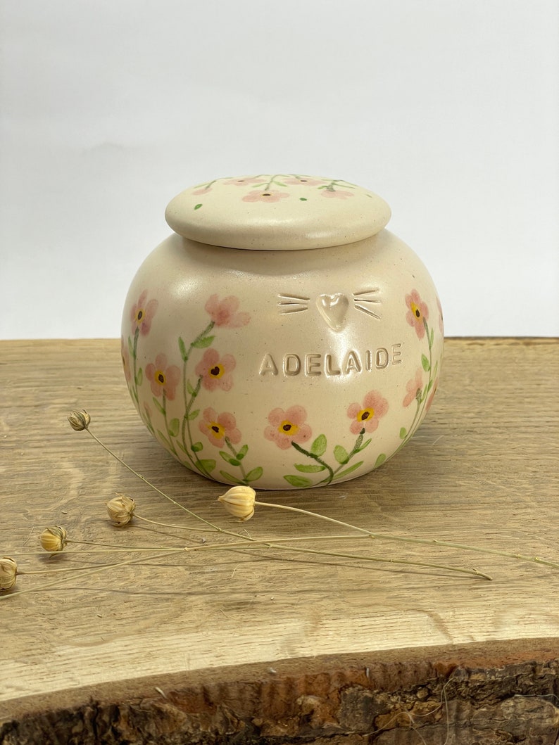 Op de afbeelding: Een kleine, cr&egrave;mekleurige keramische pot met een deksel, versierd met roze bloemen en groene bladeren. De pot heeft een kattengezicht en een hartontwerp en de naam "Adelaide" erop geschreven.