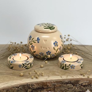 May include: A beige ceramic urn and two matching candle holders, all adorned with blue floral designs. The urn has a lid and the word "ZULA" on the front. The candle holders each hold a lit tea light.