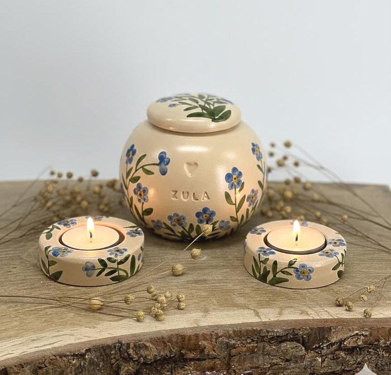 May include: A beige ceramic urn and two matching candle holders, all adorned with blue floral designs. The urn has a lid and the word "ZULA" on the front. The candle holders each hold a lit tea light.